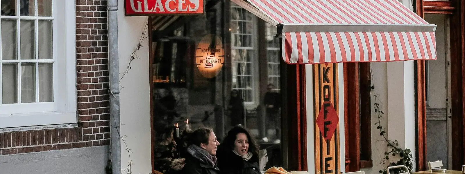 Two people at a cafe table under a red and white striped awning on a brick street